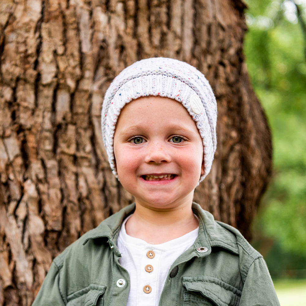 Smiling child wearing the Speckle Byron Beanie beside a tree outdoors