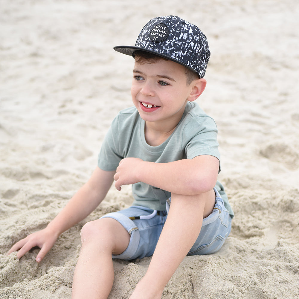 A young boy sitting on the sand, wearing a monochrome alphabet print cap with a black PU leather peak. The cap has a "Little Renegade Company" logo on the front, adding a trendy touch to his casual outfit.