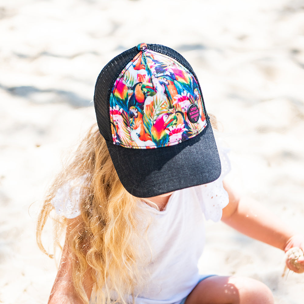 Close-up of a child wearing the Flock Trucker Cap at the beach, showing its rainbow bird print and black curved peak