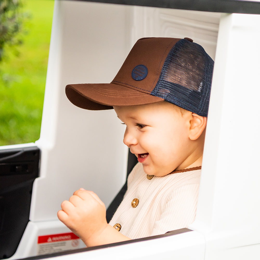 Toddler wearing the Chocolate Trucker Cap while sitting in a white toy truck, showing the brown front, curved peak and navy mesh side