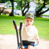 Young child wearing the forest green Hunter Trucker Cap while playing outdoors with a scooter