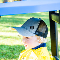 Child wearing the Shadow Trucker Cap outdoors in a yellow top, showing the grey front and black mesh side and back